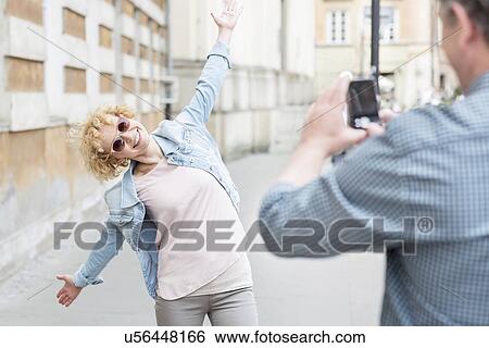 Man photographing playful woman standing with arms outstretched on city street View Large Photo Image Stock Photograph - Man photographing playful woman standing with arms outstretched on city street. Fotosearch