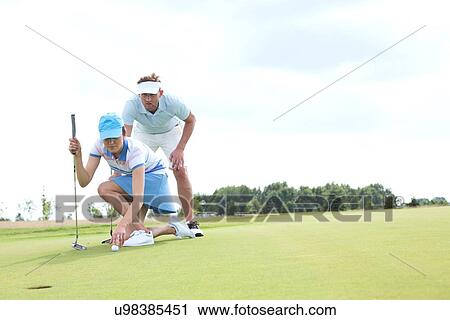 Stock Image - Man with woman aiming ball on golf course against sky. Fotosearch