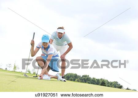 Stock Image - Mid-adult man looking at woman aiming ball on golf course. Fotosearch