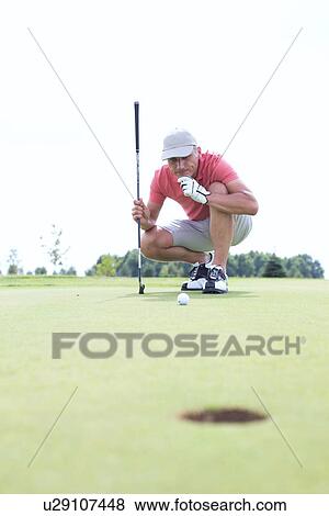 Stock Photo - Middle-aged man looking at ball while crouching on golf course. Fotosearch