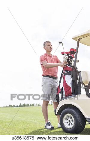 Stock Photography - Middle-aged man standing by cart at golf course. Fotosearch