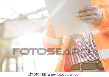 Stock Photo - Midsection supervisor holding clipboard at construction site on sunny day. Fotosearch