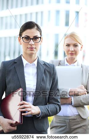 Portrait of confident businesswoman holding folder with colleague in background View Large Photo Image Picture - Portrait of confident businesswoman holding folder with colleague in background. Fotosearch