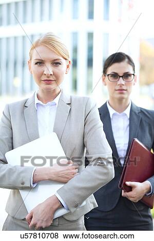 Stock Photo - Portrait of confident businesswoman holding laptop with colleague in background. Fotosearch