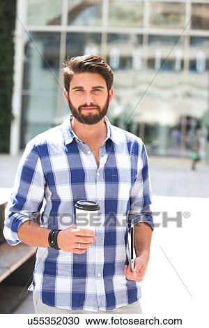 Stock Image - Portrait of confident man looking away while holding disposable cup and tablet PC in city. Fotosearch