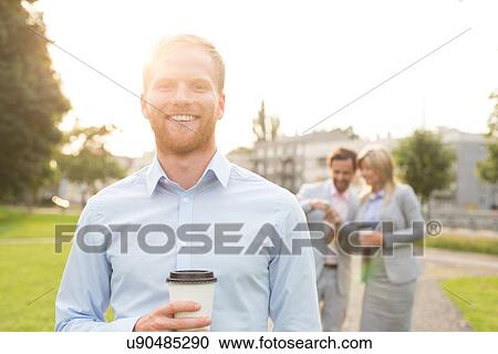 Portrait of happy businessman holding disposable cup with colleagues standing in background View Large Photo Image Stock Image - Portrait of happy businessman holding disposable cup with colleagues standing in background. Fotosearch