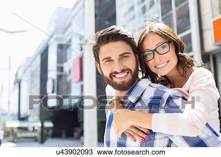 Stock Image - Portrait of happy man giving piggyback ride to woman in city. Fotosearch