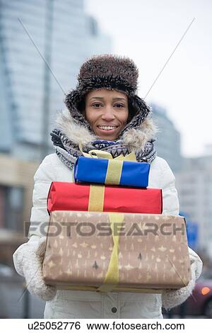 Stock Photograph - Portrait of happy woman carrying stacked gifts during winter. Fotosearch