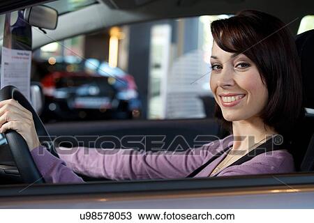 Stock Image - Portrait of young woman sitting in driver's seat at car dealersh. Fotosearch