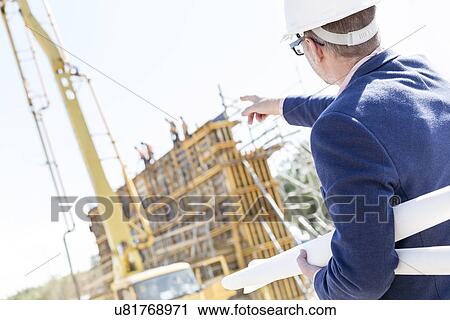 Rear view of architect holding blueprints while pointing at construction site View Large Photo Image Stock Image - Rear view of architect holding blueprints while pointing at construction site. Fotosearch