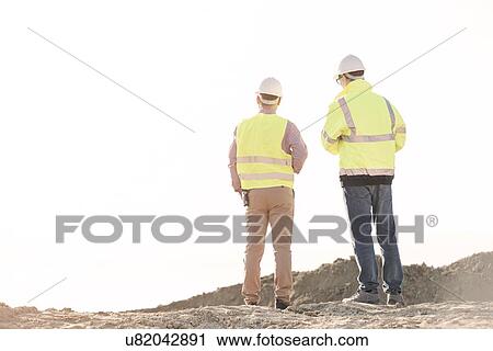 Stock Image - Rear view of architects standing at construction site against clear sky. Fotosearch