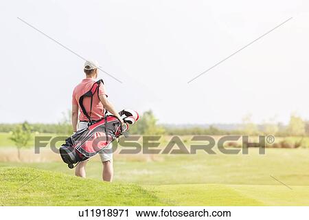 Stock Image - Rear view of man carrying golf club bag while walking at course. Fotosearch
