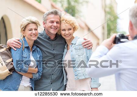Stock Image - Rear view of man photographing male and female friends in city. Fotosearch