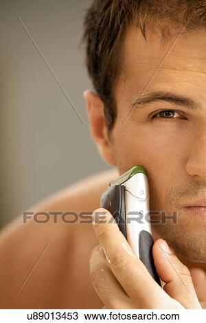 Stock Image - Reflection of young man in mirror shaving with electric shaver. Fotosearch
