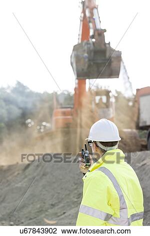 Stock Image - Side view of engineer using walkie-talkie at construction site. Fotosearch