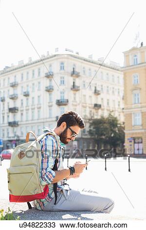 Stock Image - Side view of man with backpack using cell phone in city on sunny day. Fotosearch