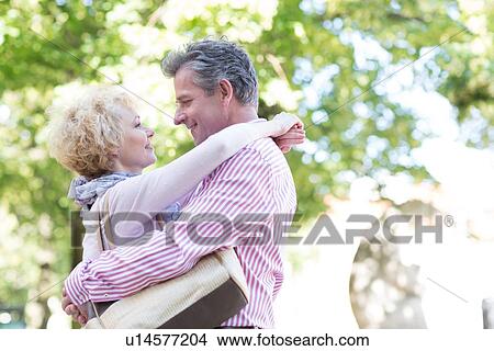 Picture - Side view of middle-aged couple embracing while looking at each other in park. Fotosearch