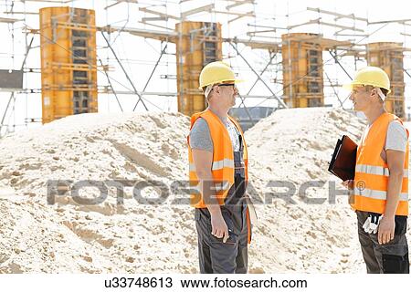 Stock Image - Side view of supervisors discussing at construction site. Fotosearch