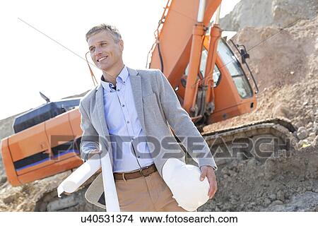 Smiling architect looking away while holding blueprints and hardhat at construction site View Large Photo Image Picture - Smiling architect looking away while holding blueprints and hardhat at construction site. Fotosearch