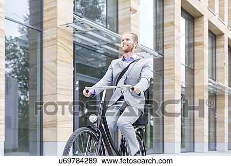 Stock Photo - Smiling businessman riding bicycle outside building. Fotosearch