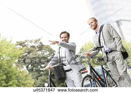 Stock Photo - Smiling businessman with bicycle showing something to colleague on street. Fotosearch