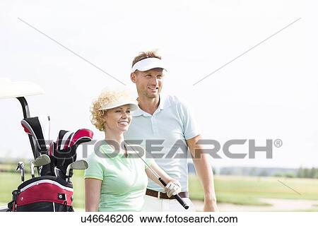 Stock Photograph - Smiling golfers standing at golf course against clear sky. Fotosearch