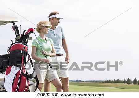 Stock Image - Smiling man and woman standing at golf course against clear sky. Fotosearch
