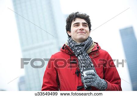 Stock Photo - Smiling man in warm clothing looking away while holding disposable cup outdoors. Fotosearch