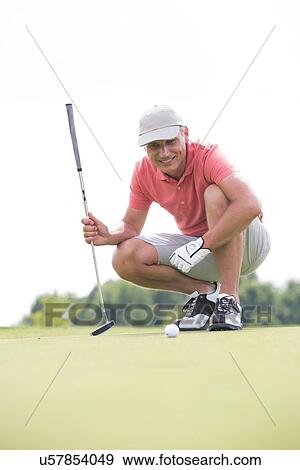 Stock Photo - Smiling middle-aged man looking at ball while crouching on golf course. Fotosearch