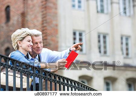 Stock Photo - Smiling middle-aged man showing something to woman with guidebook in city. Fotosearch