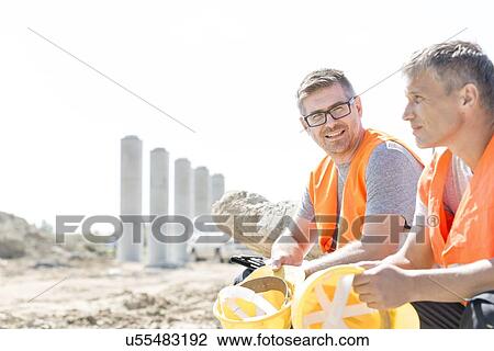 Stock Image - Smiling supervisor sitting with colleague at construction site. Fotosearch