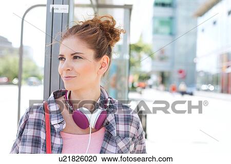 Stock Image - Smiling woman looking away while waiting at bus stop. Fotosearch