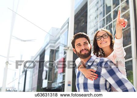 Stock Photo - Smiling woman pointing away while enjoying piggyback ride on man in city. Fotosearch