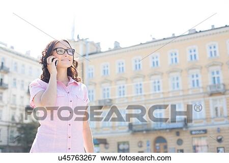 Stock Photo - Smiling young woman answering smart phone against building on sunny day. Fotosearch