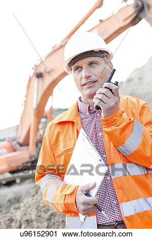 Stock Image - Supervisor holding clipboard while using walkie-talkie at construction site. Fotosearch