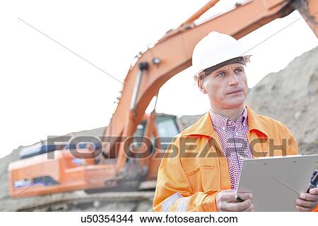 Picture - Supervisor looking away while holding clipboard at construction site. Fotosearch