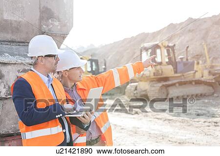 Stock Image - Supervisor showing something to coworker holding laptop at construction site. Fotosearch