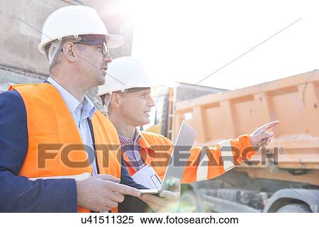 Stock Photography - Supervisor showing something to colleague holding laptop at construction site. Fotosearch