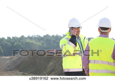 Stock Image - Supervisor using mobile phone while standing with colleague at construction site against clear sky. Fotosearch