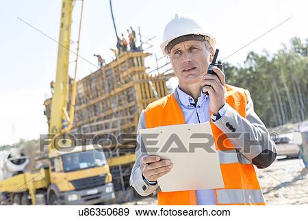 Stock Photo - Supervisor using walkie-talkie while holding clipboard at construction site. Fotosearch