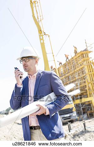 Supervisor using walkie-talkie while holding blueprints at construction site View Large Photo Image Stock Photograph - Supervisor using walkie-talkie while holding blueprints at construction site. Fotosearch