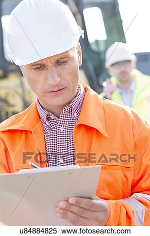 Supervisor writing on clipboard at construction site with colleague in background View Large Photo Image Stock Photography - Supervisor writing on clipboard at construction site with colleague in background. Fotosearch