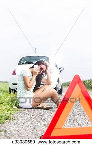 Tensed woman using cell phone while sitting on country road by broken down car View Large Photo Image Stock Photo - Tensed woman using cell phone while sitting on country road by broken down car. Fotosearch
