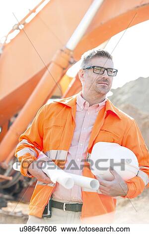 Thoughtful architect holding hardhat and blueprints at construction site View Large Photo Image Stock Photograph - Thoughtful architect holding hardhat and blueprints at construction site. Fotosearch