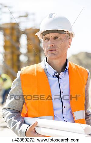 Stock Image - Thoughtful architect looking away while holding blueprints at construction site. Fotosearch
