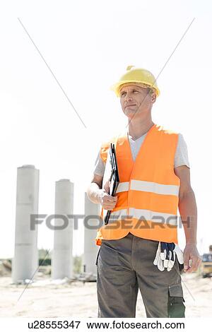 Stock Photo - Thoughtful architect looking away while holding clipboard at construction site. Fotosearch