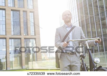 Thoughtful businessman standing with bicycle outside building View Large Photo Image Stock Photo - Thoughtful businessman standing with bicycle outside building. Fotosearch