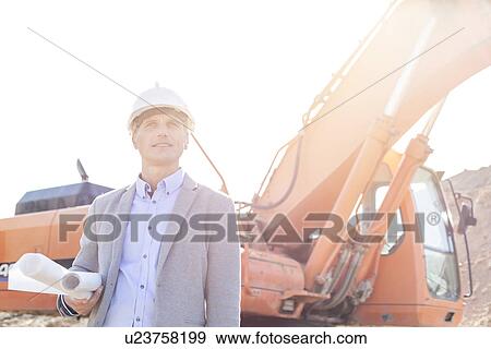 Stock Photo - Thoughtful engineer looking away while holding blueprints by bulldozer at construction site. Fotosearch