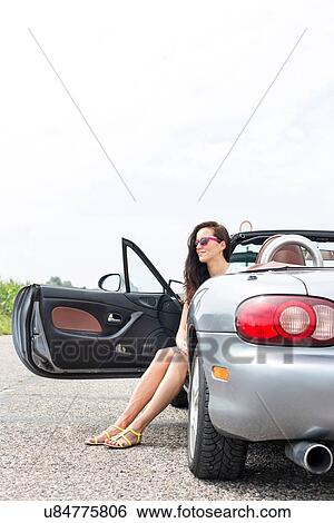 Stock Photograph - Thoughtful woman sitting in convertible on country road against clear sky. Fotosearch