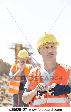 Thoughtful worker standing at construction site with colleague in background View Large Photo Image Stock Photo - Thoughtful worker standing at construction site with colleague in background. Fotosearch